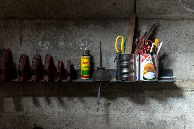 A garage wall with a shelf, cans of tools, oil, and clamps