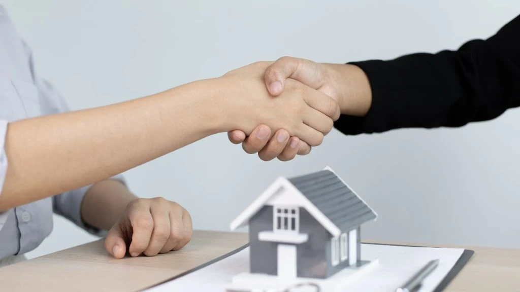Two people shaking hands over a small replica of a house