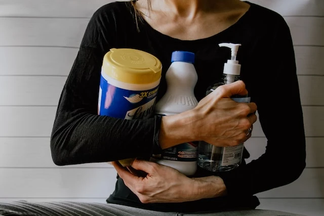 Cropped image of a person in a balck shirt holding a container of wipes, a bottle of bleach, and a bottle of hand sanitizer in their arms