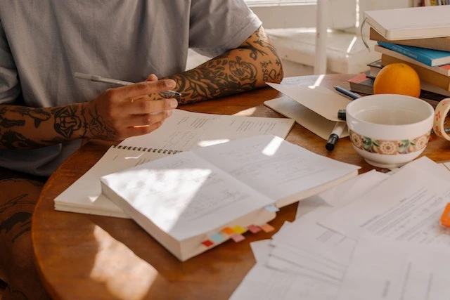 Cropped image of a person with tattoos on their arms studying a book and notes at a table