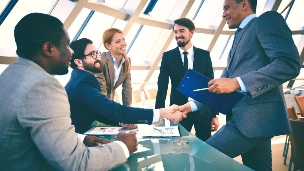 Group of five people in suits, two of the are shaking hands across a table