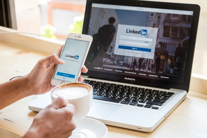 Person holding a cappuccino in one hand and their phone in another, looking at LinkedIn on their smartphone and the laptop in front of them