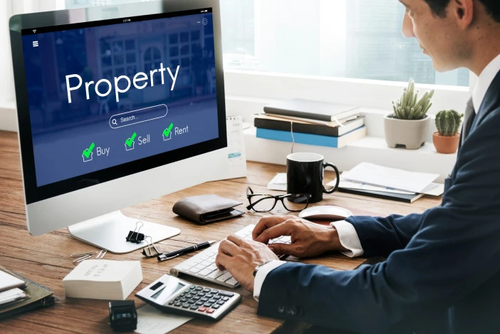 Man in a suit working at a desk, the desktop computer in front of him has a blue screen with the word “Property” on it in large letters