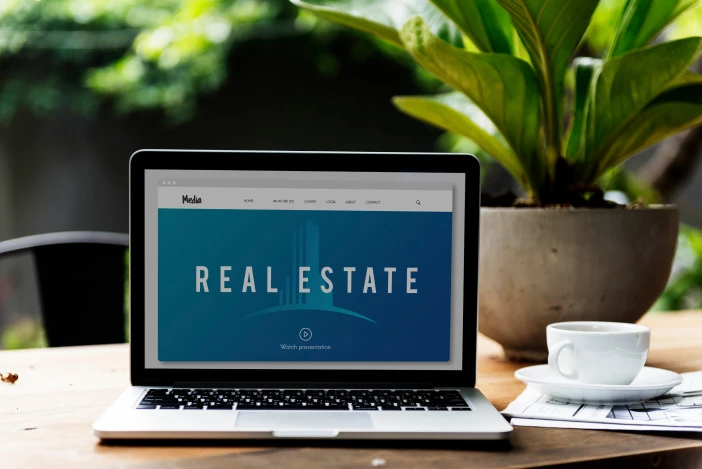Laptop displaying a real estate marketing website on an outdoor table with coffee and a plant