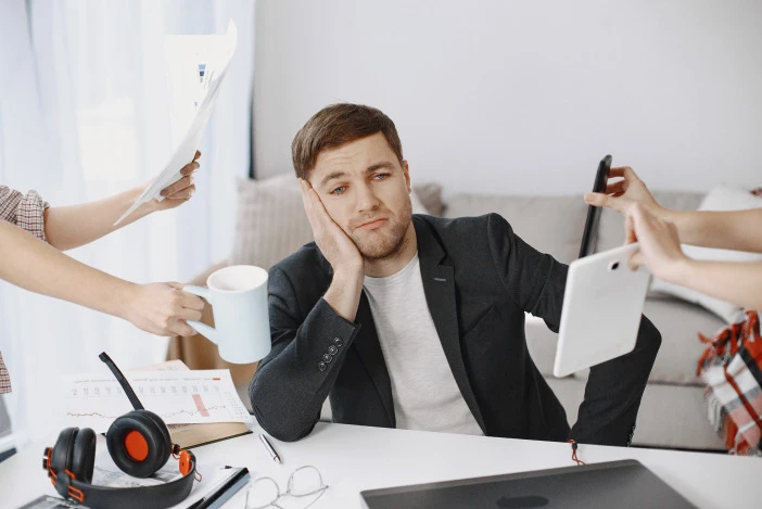 Man in a sports coat sitting at a desk, other people’s hands are coming in from the edges of the frame holding coffee cups, papers, and a tablet