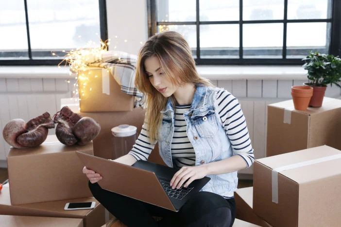 Woman sitting on the floor looking at her laptop, she’s surrounded by cardboard moving boxes