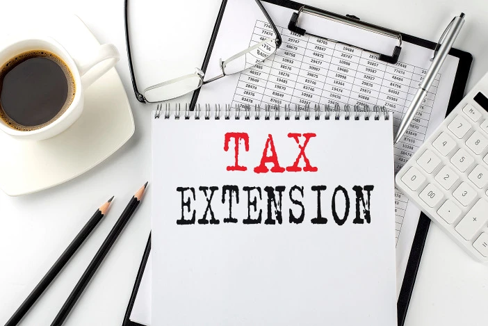 A desk with a coffee cup, glasses, pens, and a notebook that says “TAX EXTENSION” on it