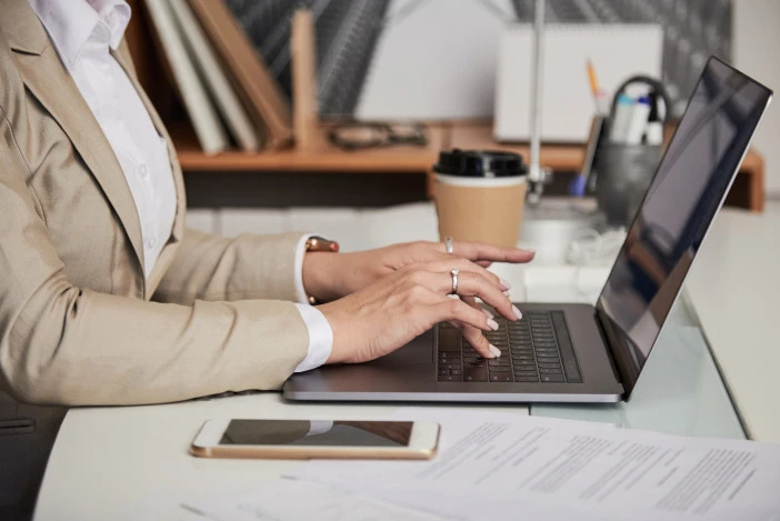 Close-up of hands typing on a laptop next to a phone and paperwork