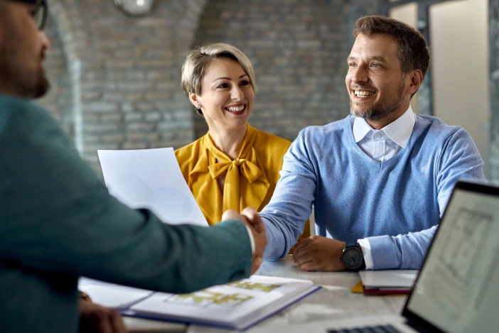 A smiling man and woman sitting across a desk from their property manager, the man is shaking the property manager’s hand