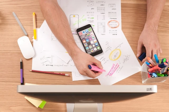 Overhead shot of someone’s arms working on a desk with paper, a phone, and a desktop computer