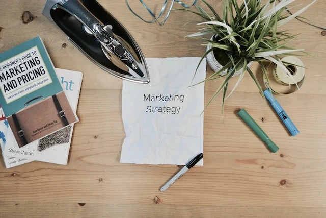 Paper that says marketing strategy on a wooden table surrounded by pens, books, a green plant, and an iron