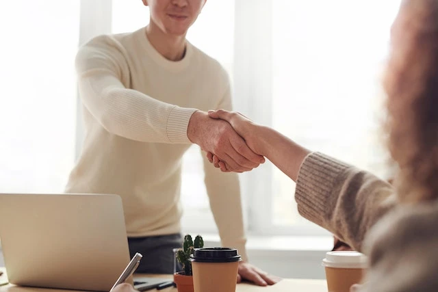 Person in a white sweater leaning across a table to shake another person’s hand