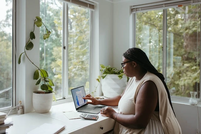 A property manager working from home at their desk in front of a window using a calculator and laptop