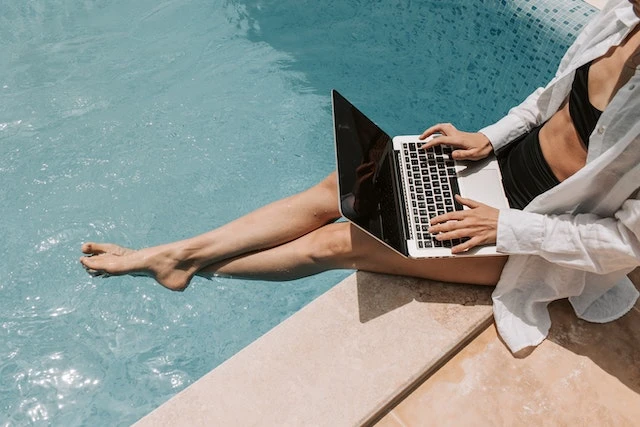 Person wearing a black two-piece bathing suit on the edge of a pool holding a laptop