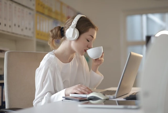 Person wearing headphones at their desk while they work on their laptop and drink a cup of tea