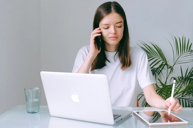 Person sitting at a desk talking on a phone while looking at a laptop and writing on a tablet