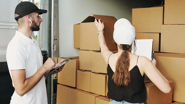 Two people wearing baseball caps looking at cardboard boxes in a moving van
