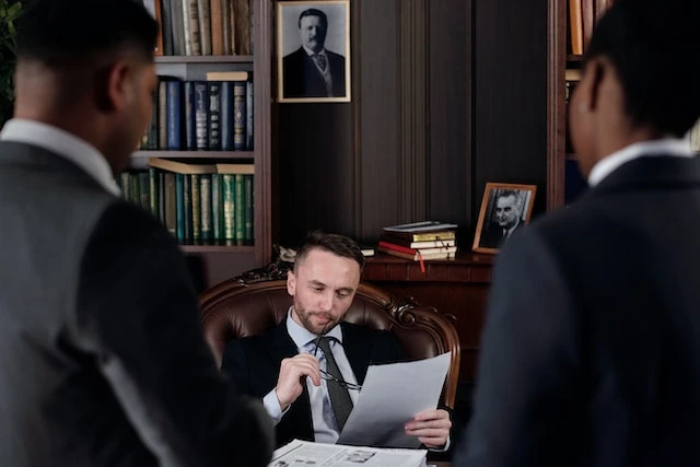 Lawyer sitting in a chair reviewing a document behind a desk, two other people in suits are standing facing him