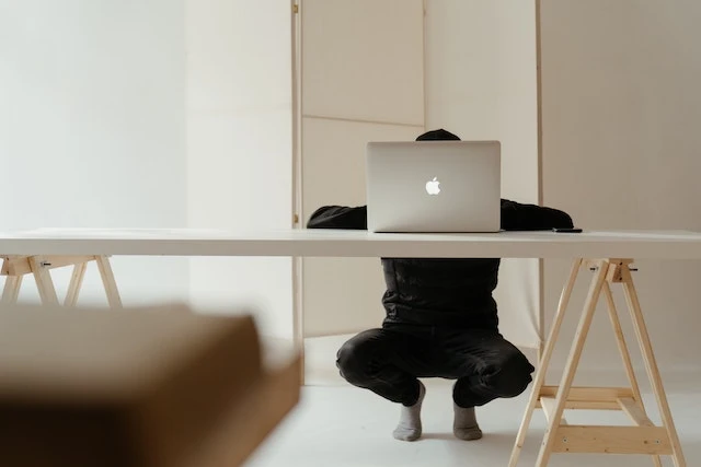 Person crouching behind a desk with a laptop covering their face