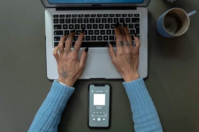 Hands typing on a laptop with a smartphone in between their arms