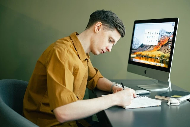 Person writing in a notebook at a desk in front of a desktop computer