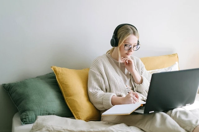 Person sitting in their bed wearing headphones and studying with a book and laptop