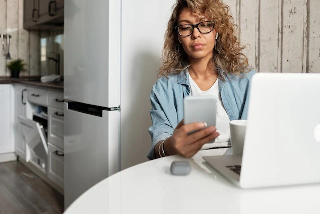 Property manager sitting in front of her laptop in her kitchen looking at the phone in her hand