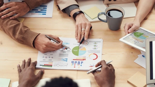 Three people’s hands holding pens and pointing at a piece of paper with colorful graphs on it