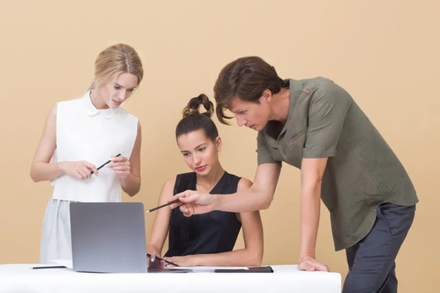 Two people looking at a laptop of a third person sitting at a desk