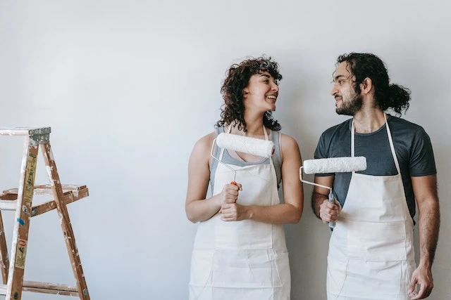 Two people wearing white aprons in a white room holding paint rollers and smilking at each other