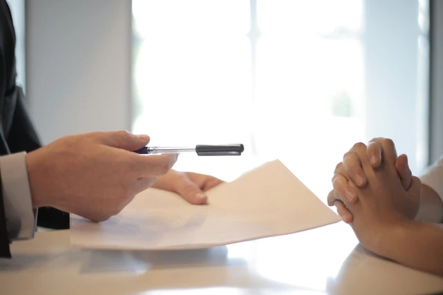 Two people’s hands across a desk, one of them is holding a document and pen