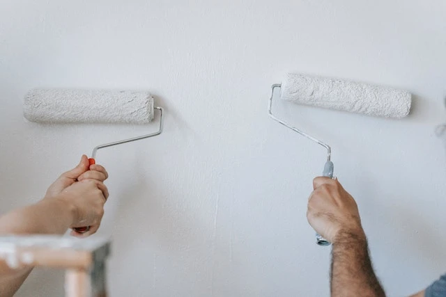 Two people’s hands using paint rollers to paint a white wall