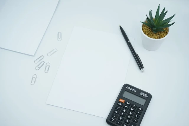 White paper on a white desk next to 6 paperclips, a black calculator, a black pen, and a small succulent