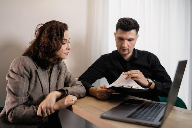 two people looking at a clipboard together at a small desk with a laptop on it