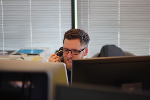 Person in an office sitting behind a desktop computer talking on a landline phone