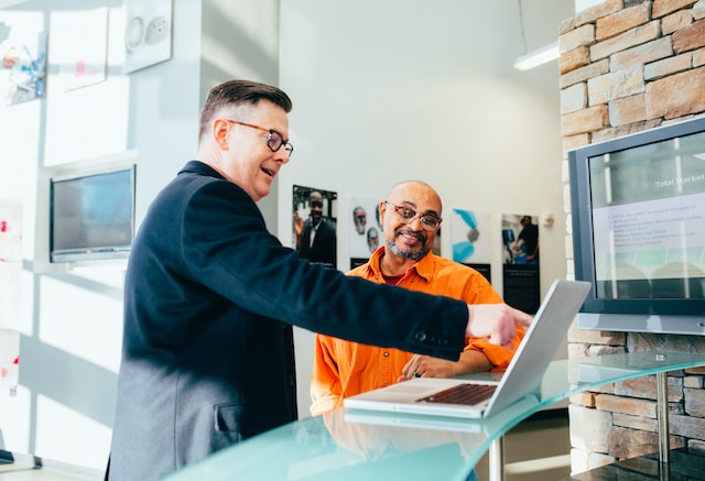 Two people standing at a desk, one of them is pointing at a laptop