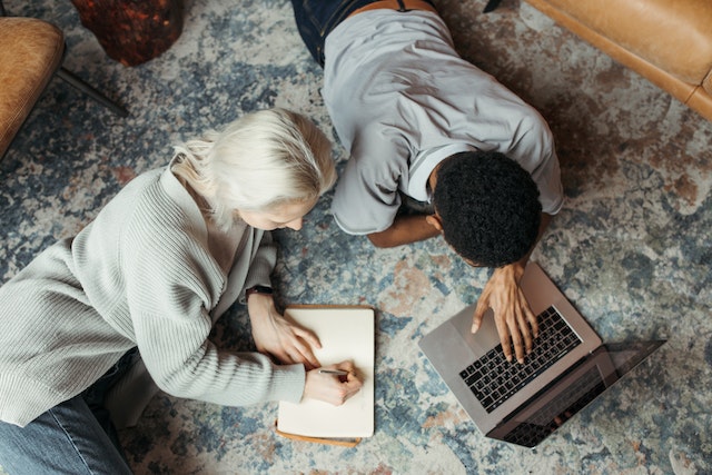 Overhead of two people working on a carpet, one is writing in a notebook and the other is working on a laptop