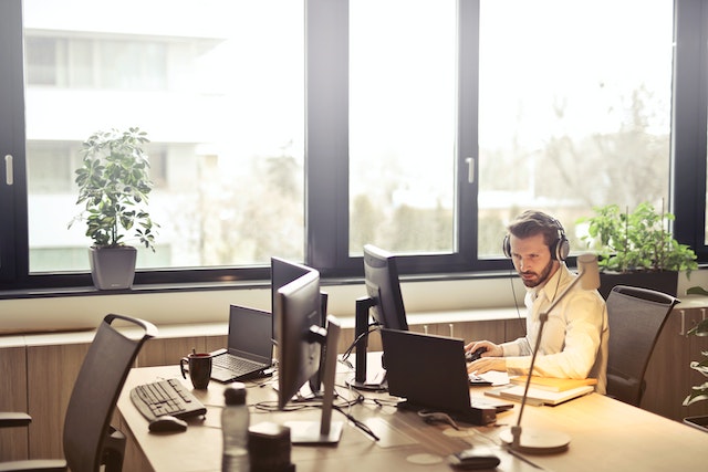 Man working in an office on multiple computers while wearing headphones