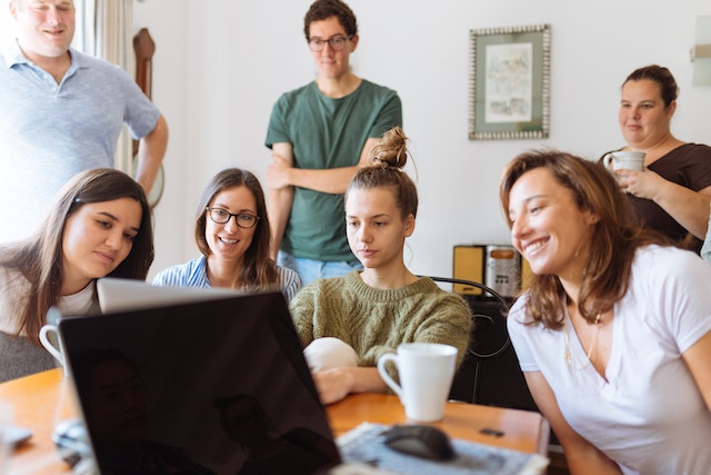 Group of seven people sitting around a table all looking at the same laptop