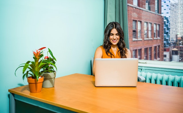 Smiling woman working on a laptop at a table in front of a turquoise wall and a window