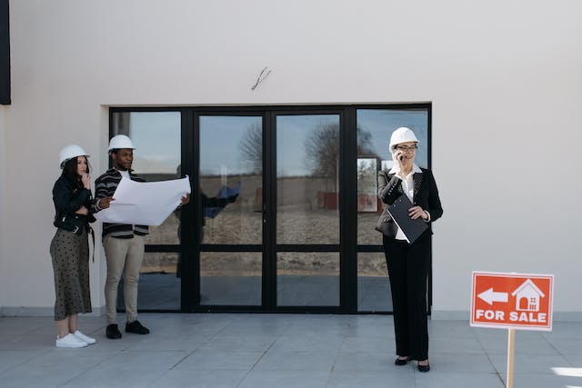 Three people in hard hats in front of a property for sale looking at blueprints