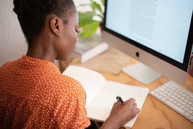 Woman in an orange shirt working on a desktop computer and writing in a notebook