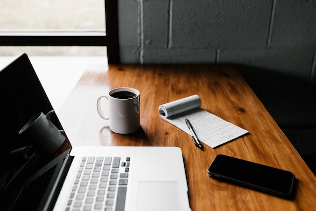 Wooden desk set up with a laptop, coffee cup, notepad and pen, and smartphone