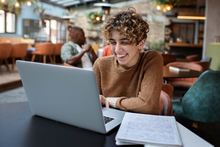 Smiling person with short curly hair taking notes and working on their laptop in a cafe