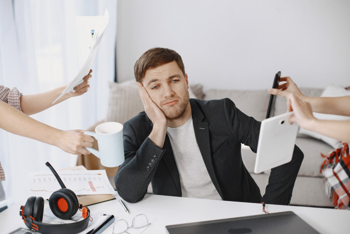 Man in a sports coat sitting at a desk, other people's hands are coming in from the edges of the frame holding coffee cups, papers, and a tablet