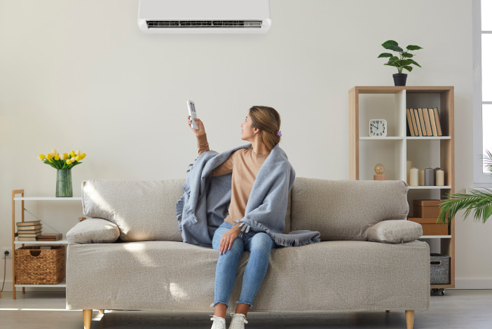 A young woman sitting on a grey couch pointing a remote at her built-in air conditioning unit