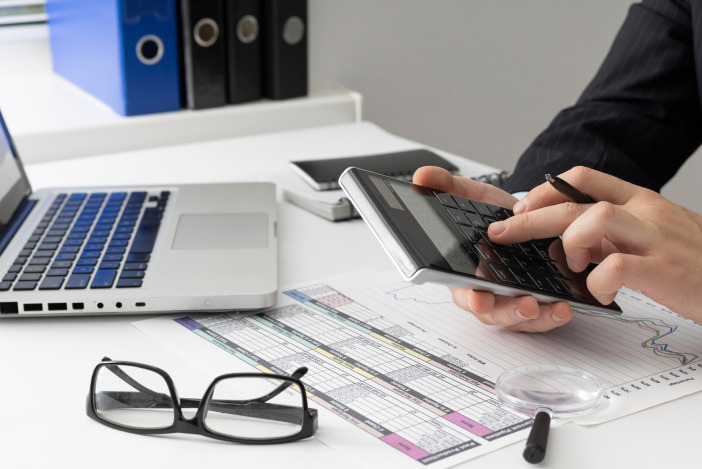 A close up of hands using a calculator, in front of a desk with papers, glasses, and a laptop