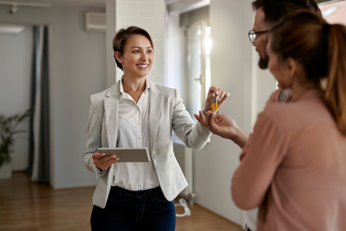 A property manager speaking to a couple, she is handing them keys with one hand and holding a tablet with the other