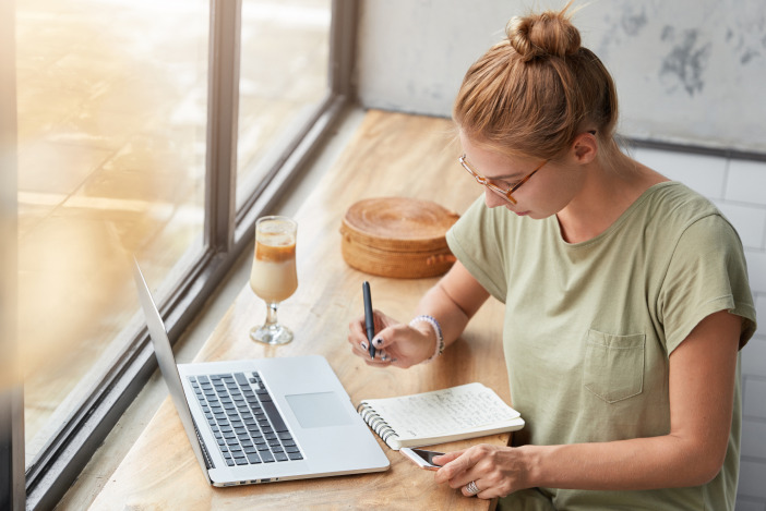 Focused woman working on her laptop and writing in a notebook in front of a large cafe window